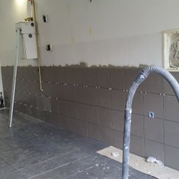 DomBud - Układanie ciemnych płytek na ścianie łazienki w trakcie prac, widoczna drabina, rura od odkurzacza i instalacja elektryczna.