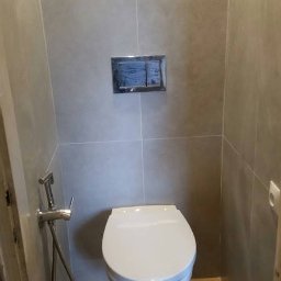 DomBud - Nowoczesna toaleta podwieszana z chromowanym przyciskiem spłukującym i bidetką w łazience wykończonej płytkami w odcieniach szarości i drewna.