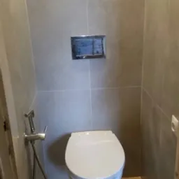 Nowoczesna toaleta podwieszana z chromowanym przyciskiem spłukującym i bidetką w łazience wykończonej płytkami w odcieniach szarości i drewna.