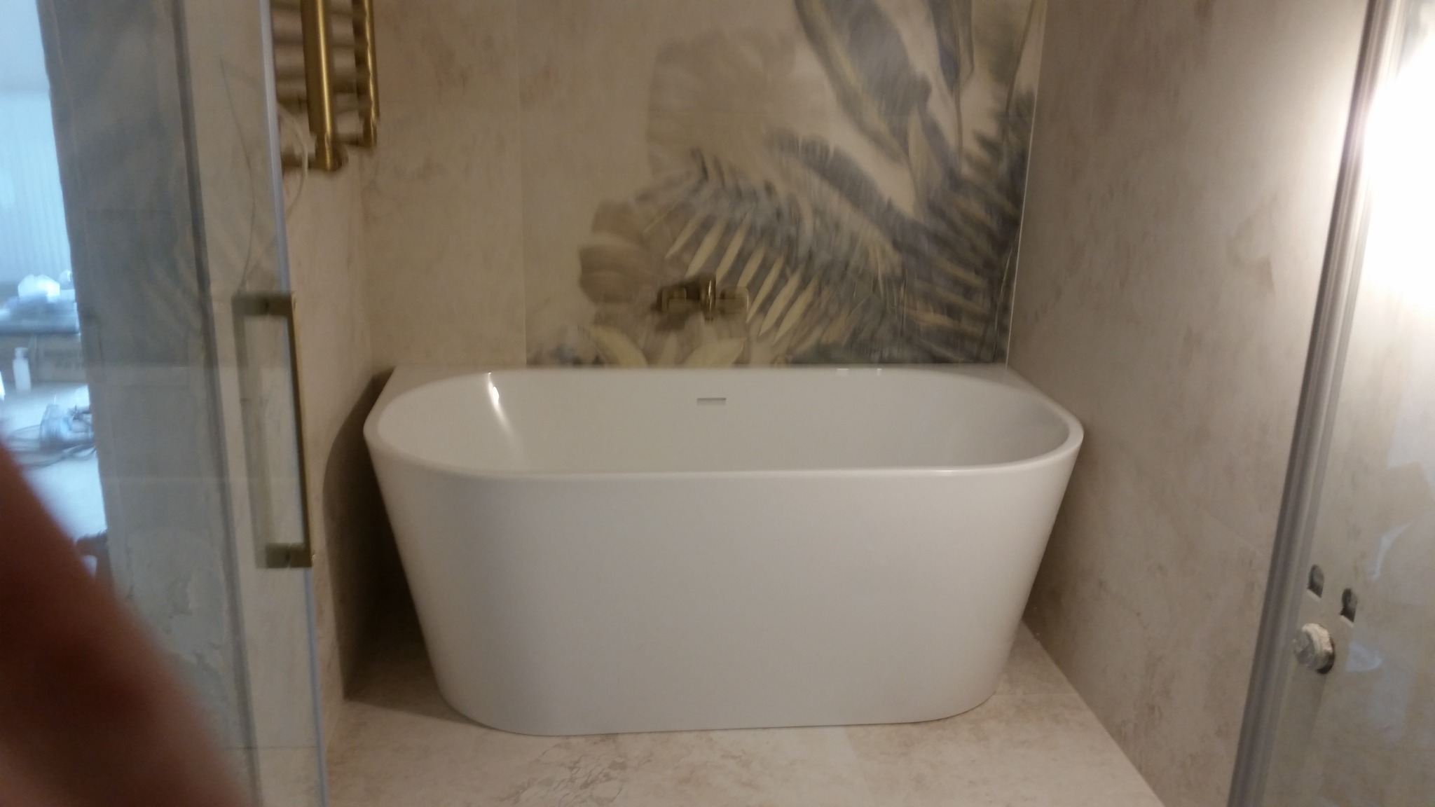 Biała, wolnostojąca wanna z baterią podtynkową na tle ściany wyłożonej płytkami z motywem liści, widok zza szklanej kabiny prysznicowej.