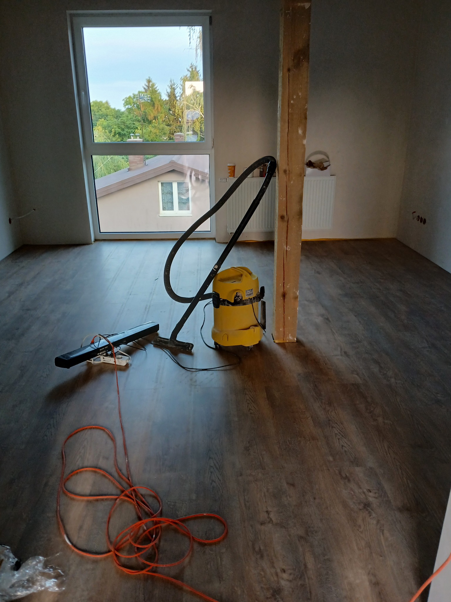 Wnętrze pokoju z nową, ciemną podłogą drewnopodobną w trakcie sprzątania, widoczny żółty odkurzacz przemysłowy, listwa przypodłogowa w trakcie montażu oraz surowy, drewniany słup konstrukcyjny.
