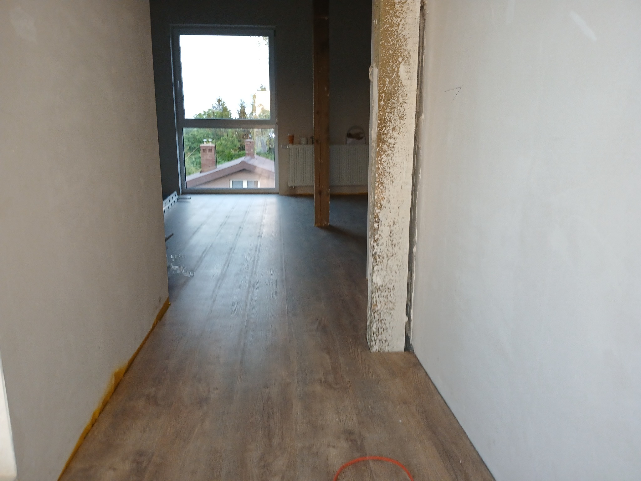 Wnętrze w trakcie prac wykończeniowych: świeżo położona podłoga laminowana, surowe ściany przygotowane do malowania, widoczna izolacja przy podłodze, okno z widokiem na zieleń.