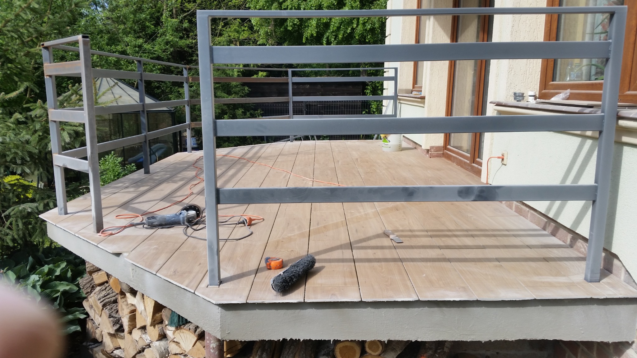 Drewniany taras w trakcie budowy z szarą, metalową balustradą, widoczne narzędzia: szlifierka kątowa, miarka i pędzel, w tle dom i stos drewna.