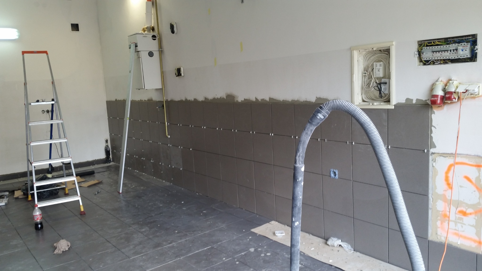 Układanie ciemnych płytek na ścianie łazienki w trakcie prac, widoczna drabina, rura od odkurzacza i instalacja elektryczna.