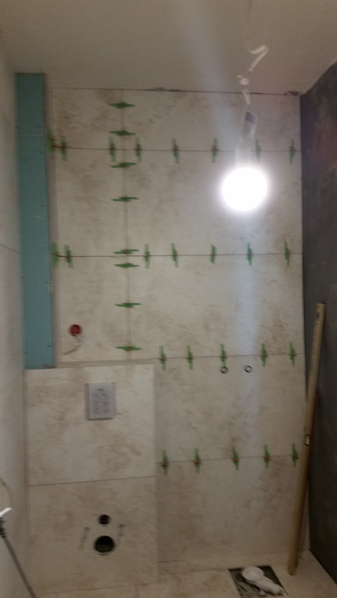 Ściana łazienki w trakcie kładzenia płytek, widoczne sznurki i krzyżyki dystansowe, fragment zabudowy geberitu, pionowy poziomnica oparta o ścianę, surowy stan.