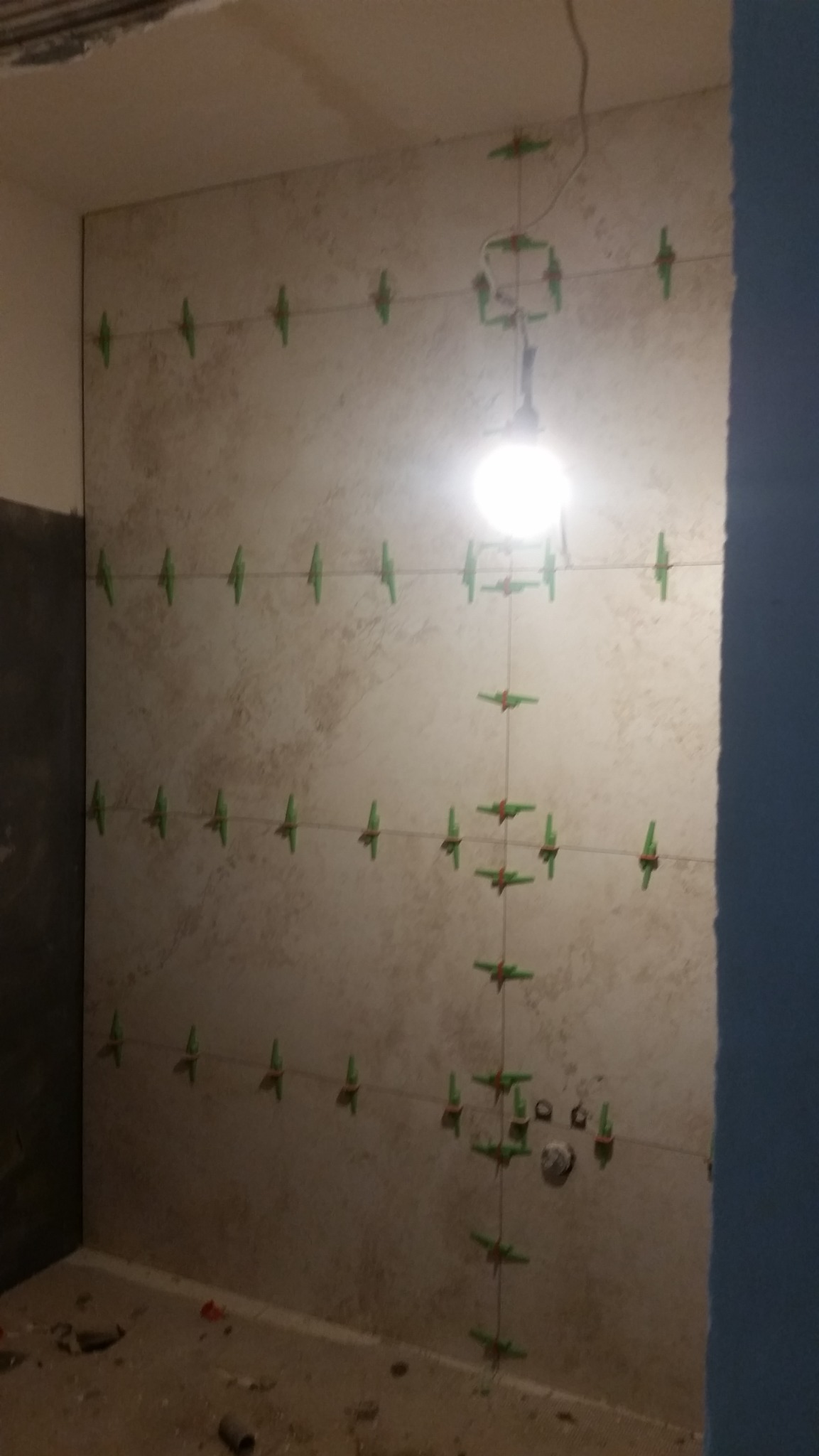 Ściana w trakcie układania płytek, widoczne zielone klipsy poziomujące, oświetlenie robocze i surowy stan pomieszczenia.