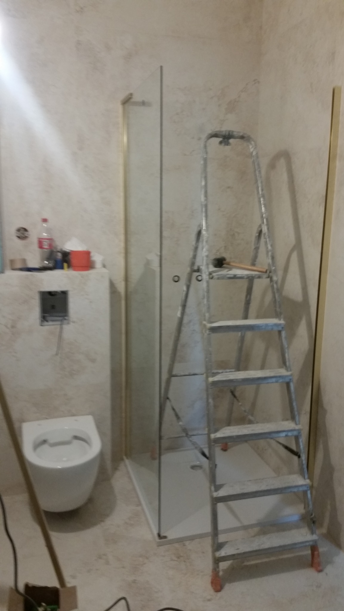 Montaż kabiny prysznicowej w łazience z zamontowaną podtynkową toaletą, widoczna drabina z młotkiem na górnym szczeblu, butelka i puszka na zabudowie stelaża.
