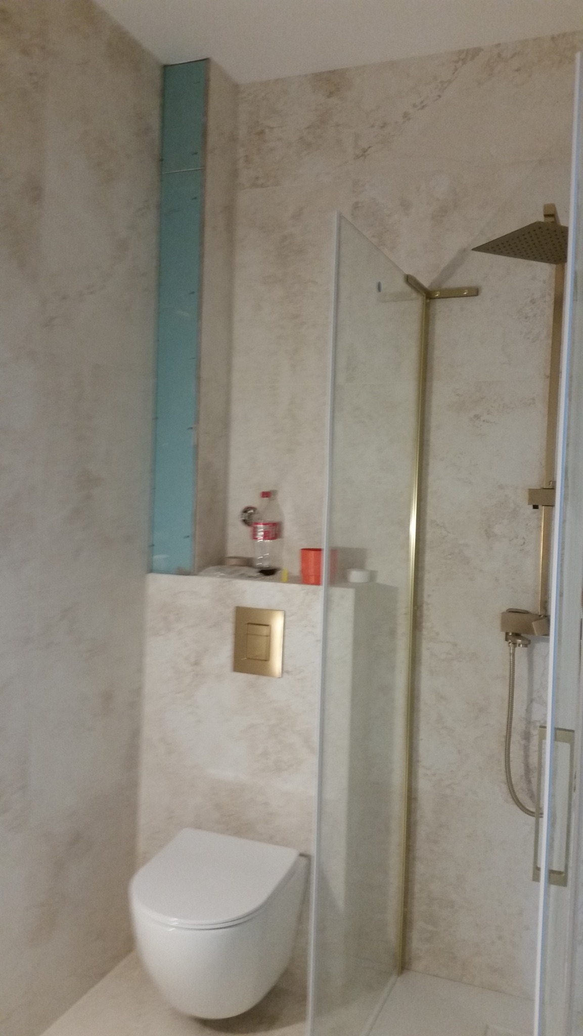 Niedokończona łazienka z zamontowaną toaletą wiszącą, kabiną prysznicową z złotymi elementami, widoczną płytą karton gips i butelką Coca-Coli na półce.