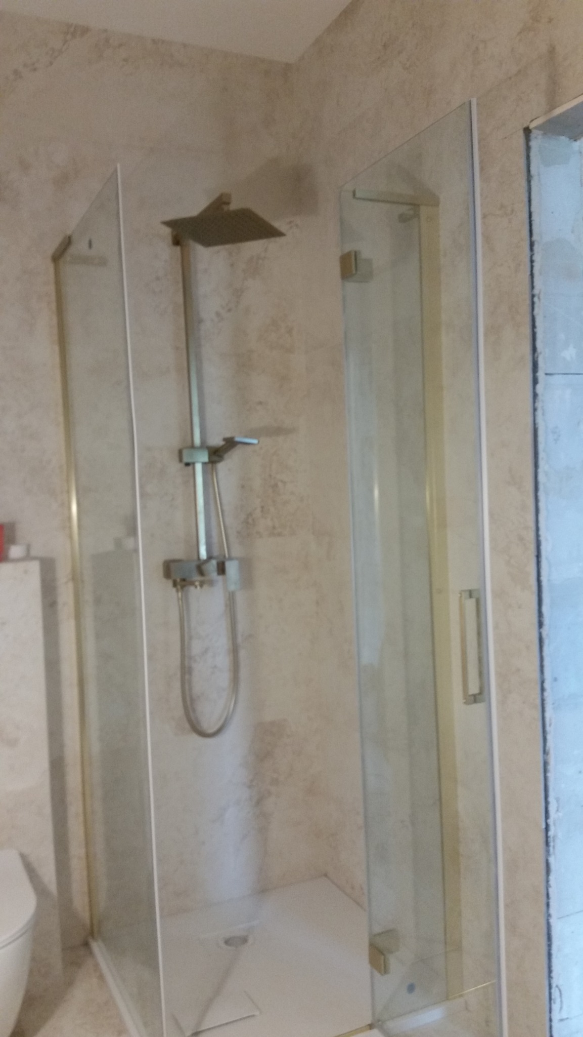 Szklana kabina prysznicowa z deszczownicą i panelem natryskowym, zamontowana w łazience z beżowymi ścianami o nieregularnej fakturze, widoczny fragment toalety po lewej stronie.