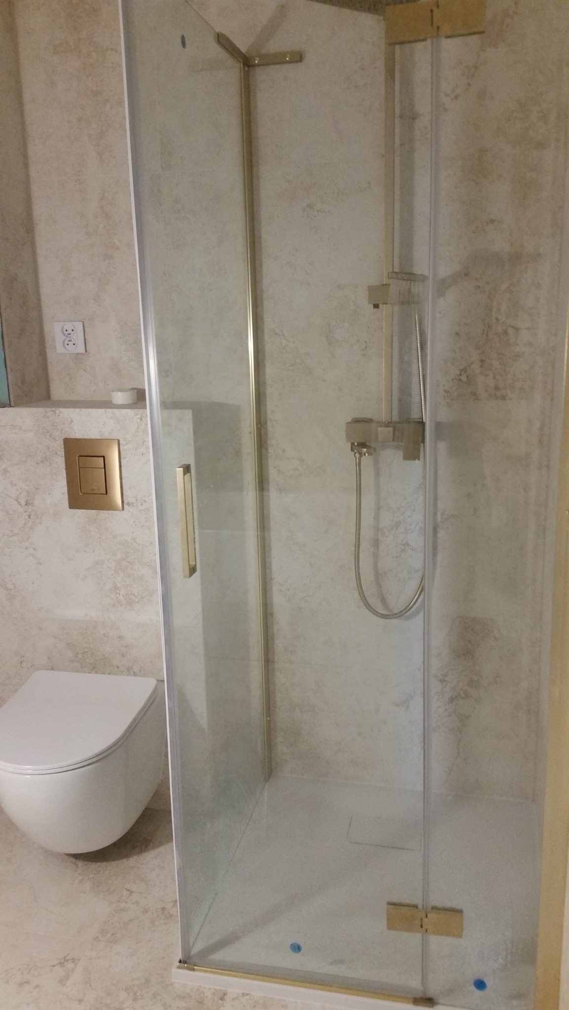 Nowoczesna kabina prysznicowa z elementami w kolorze złotym, biała wisząca toaleta i płytki imitujące kamień w łazience.