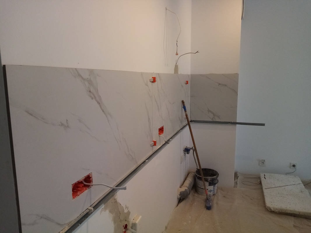 Montaż marmuropodobnych płytek ściennych w trakcie realizacji, widoczne krzyżyki dystansowe, puszki elektryczne z przewodami, wiadro i narzędzia.