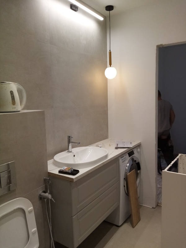 Biała umywalka wpuszczona w szafkę z szufladami, obok pralka i wisząca lampa z kulistym kloszem, widoczne płytki w odcieniach szarości i fragment toalety w trakcie montażu.