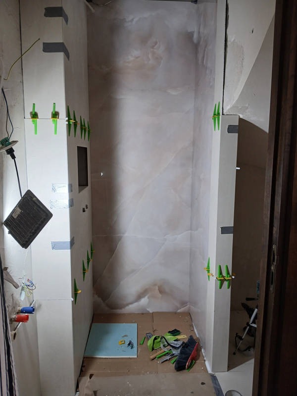 Wnętrze łazienki w trakcie prac wykończeniowych, widoczne płytki imitujące marmur na ścianach, narzędzia na podłodze oraz listwy montażowe z zielonymi krzyżykami dystansowymi na ścianach...