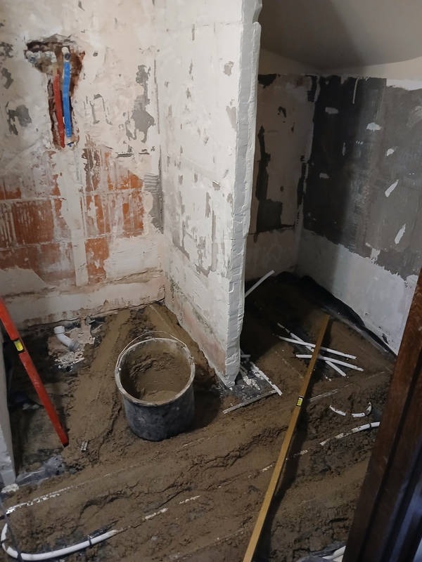 Nierówna podłoga w trakcie wyrównywania w pomieszczeniu z częściowo otynkowanymi ścianami, widoczne rury i poziomice