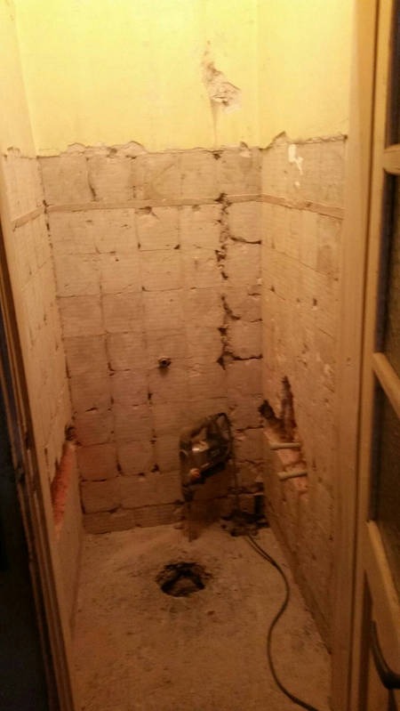 Demontaż starej glazury w małej łazience, widoczne ślady po płytkach na ścianach, odsłonięty mur, otwór w podłodze, narzędzie do cięcia płytek.