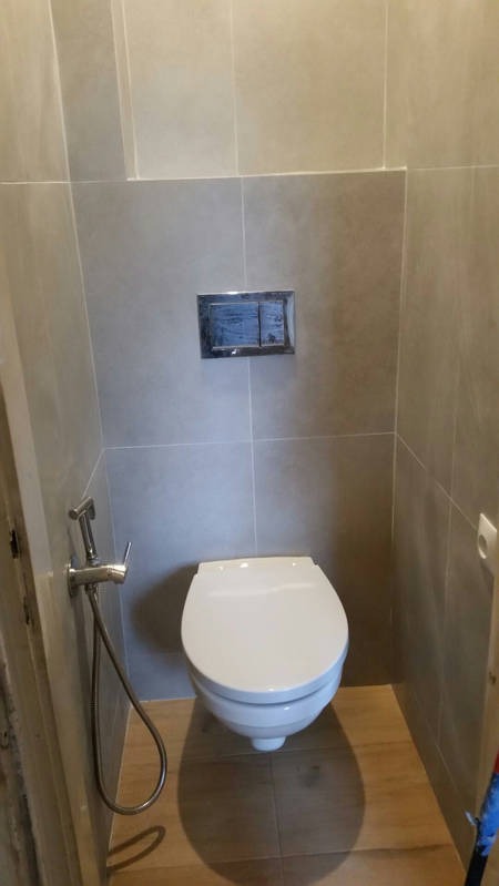 Nowoczesna toaleta podwieszana z chromowanym przyciskiem spłukującym i bidetką w łazience wykończonej płytkami w odcieniach szarości i drewna.