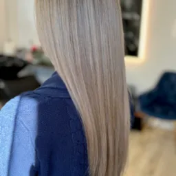 Długie, proste włosy w odcieniach blondu, z widoczną strukturą i refleksami, opadające na granatowy sweter w warkocze, w tle rozmyte wnętrze salonu.