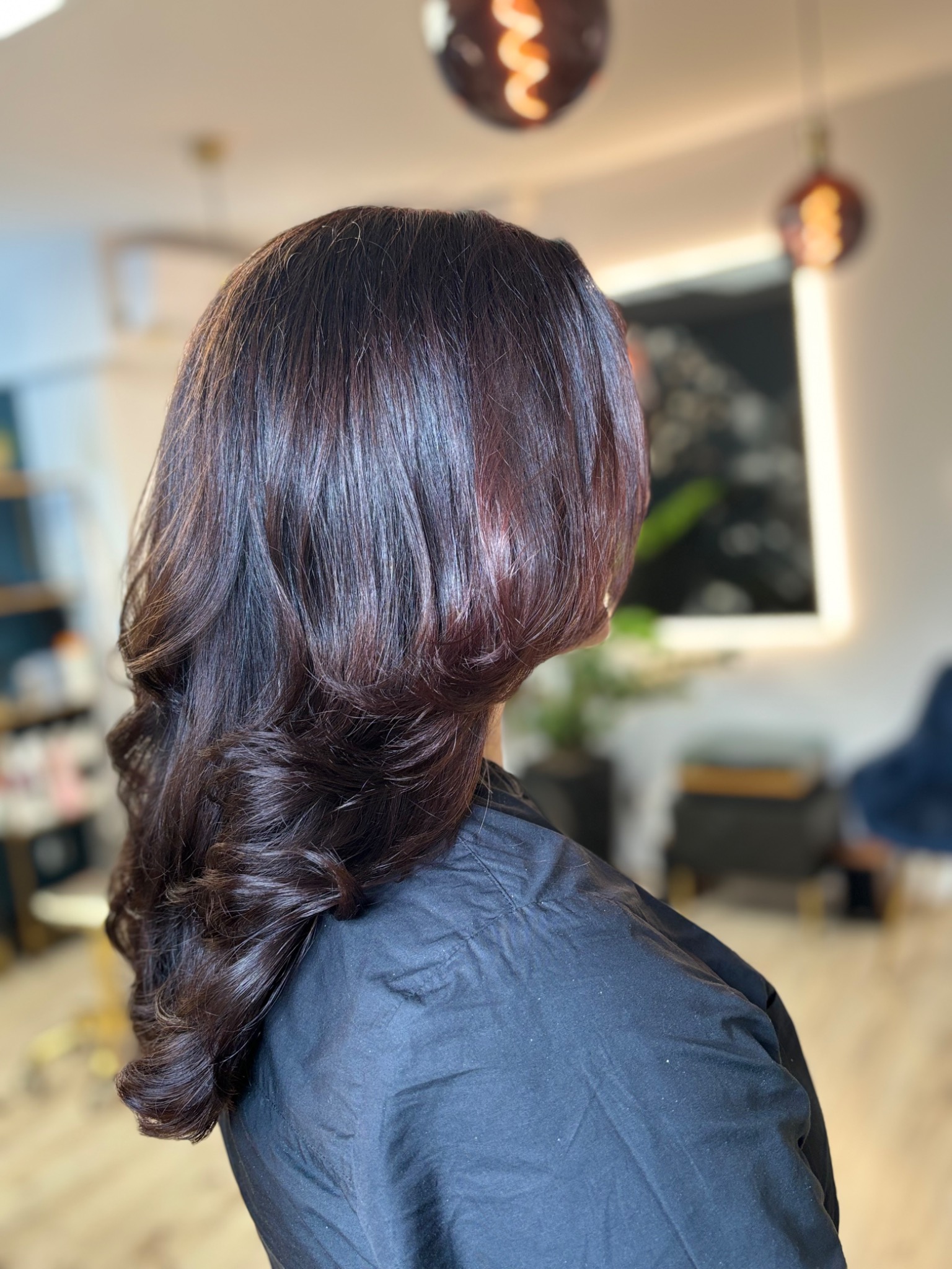 Ciemne, pofalowane włosy z subtelnymi refleksami, widoczne od tyłu, z rozmyciem tła i dekoracyjnymi lampami wiszącymi.