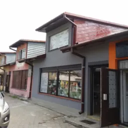 Fasada szeregu budynków z różnymi kolorami i materiałami wykończeniowymi, w tym szarym tynkiem, pomarańczowymi elementami i drewnianą okładziną, z szyldem reklamowym na jednym z budynków...