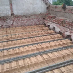 Przygotowanie podłoża pod wylewkę na zewnątrz budynku, widoczne płyty z OSB i siatka zbrojeniowa, w tle fragment ceglanej ściany z ubytkami tynku i butelka wody.