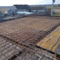 Zbrojenie stropu z siatki stalowej na żółtym szalunku, widok z góry na budowę domu z wiejską zabudową w tle.