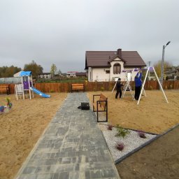 Wojcik - Plac zabaw z huśtawkami i zjeżdżalnią, na piaszczystym podłożu, z ławeczkami i ścieżką z kostki, w tle dom jednorodzinny i drewniane ogrodzenie.
