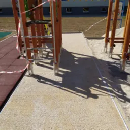 Montaż placu zabaw: drewniana konstrukcja z drabinkami na żwirowym podłożu, obok gumowa nawierzchnia, teren zabezpieczony taśmą ostrzegawczą.