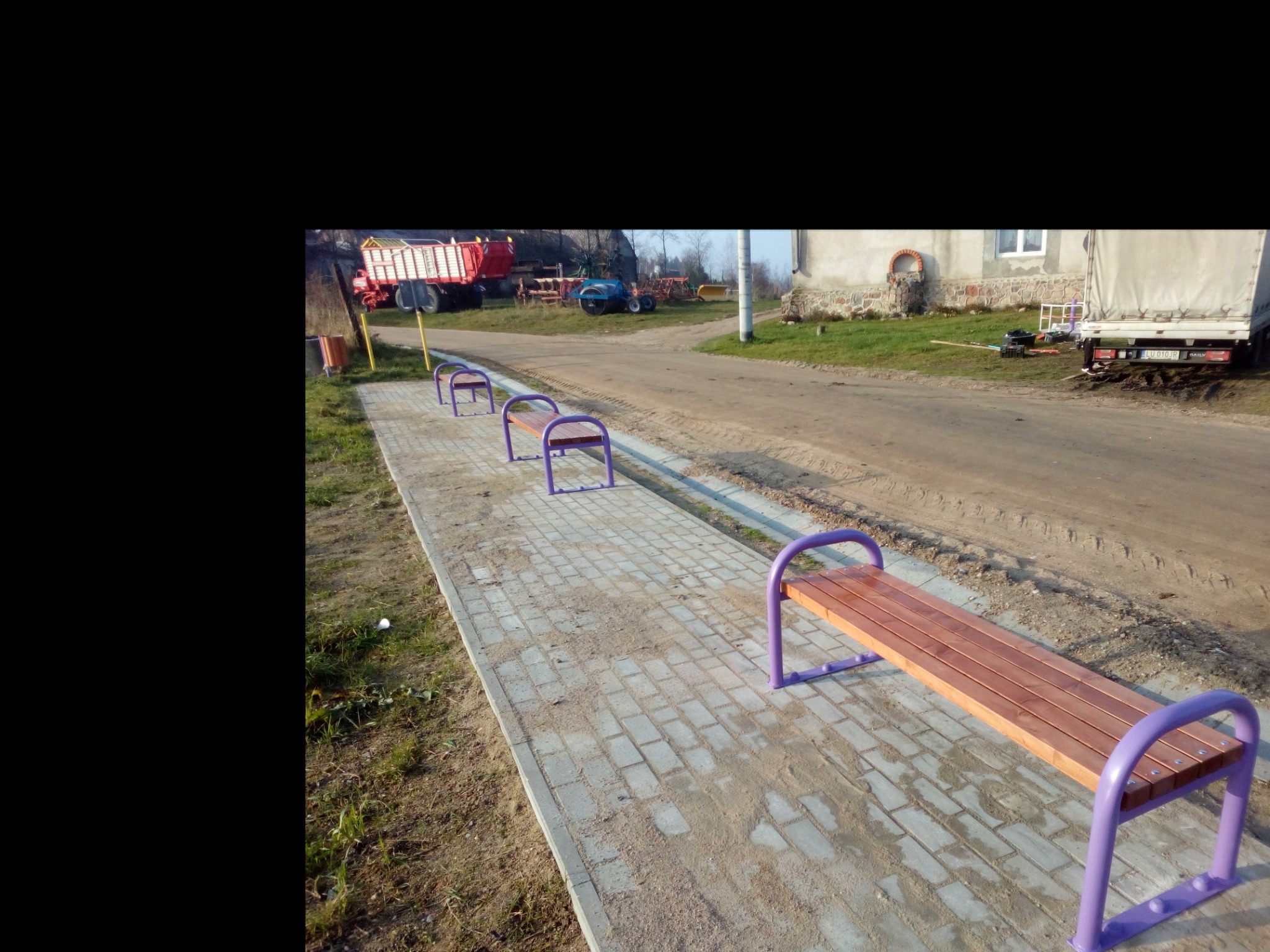 Ustawione w rzędzie drewniane ławki parkowe z fioletowymi metalowymi elementami, ułożone na chodniku z kostki, w tle wiejskie zabudowania i maszyny rolnicze.
