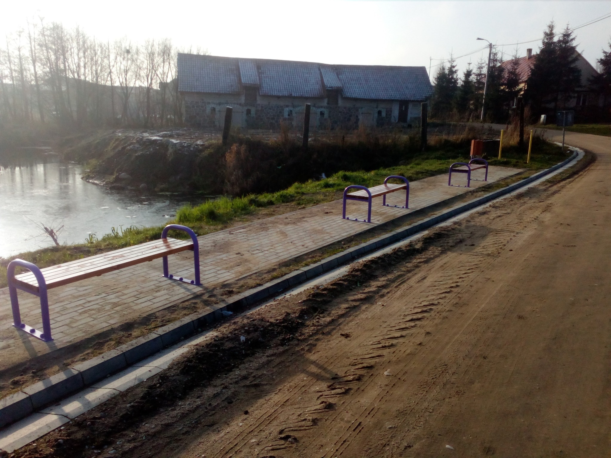 Trzy nowe ławki z drewnianym siedziskiem i fioletowymi metalowymi elementami ustawione wzdłuż chodnika z kostki brukowej obok stawu w wiejskim otoczeniu.