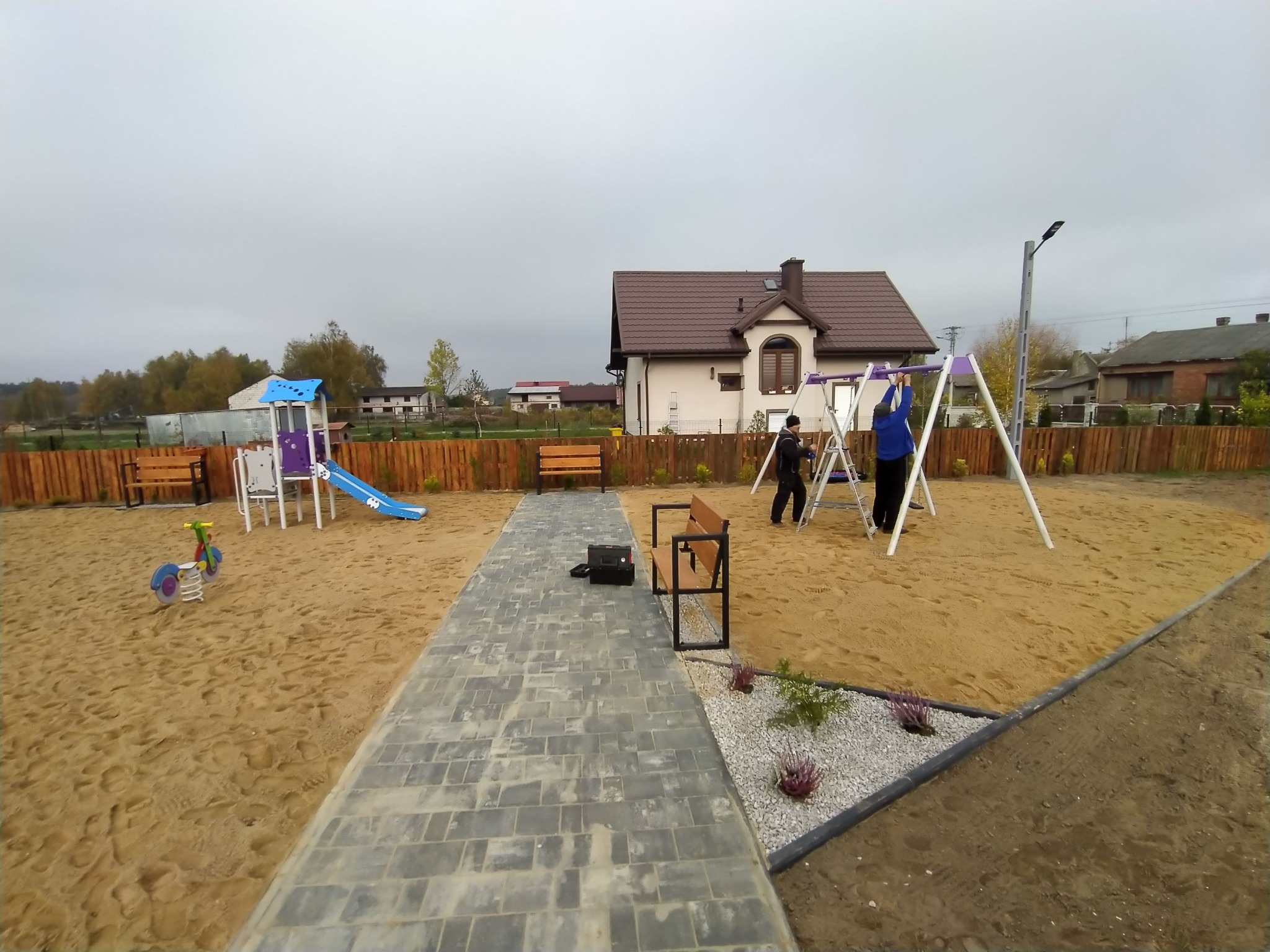 Plac zabaw z huśtawkami i zjeżdżalnią, na piaszczystym podłożu, z ławeczkami i ścieżką z kostki, w tle dom jednorodzinny i drewniane ogrodzenie.