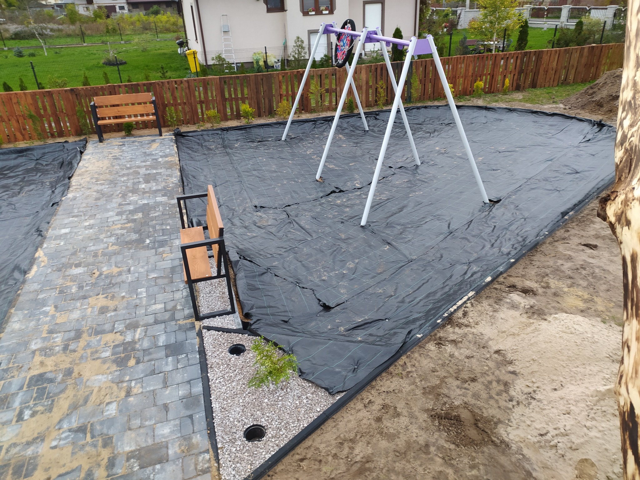 Przygotowanie terenu pod plac zabaw: rozłożona czarna agrowłóknina na ziemi, w tle huśtawka, obok ścieżka z kostki brukowej i ławka.