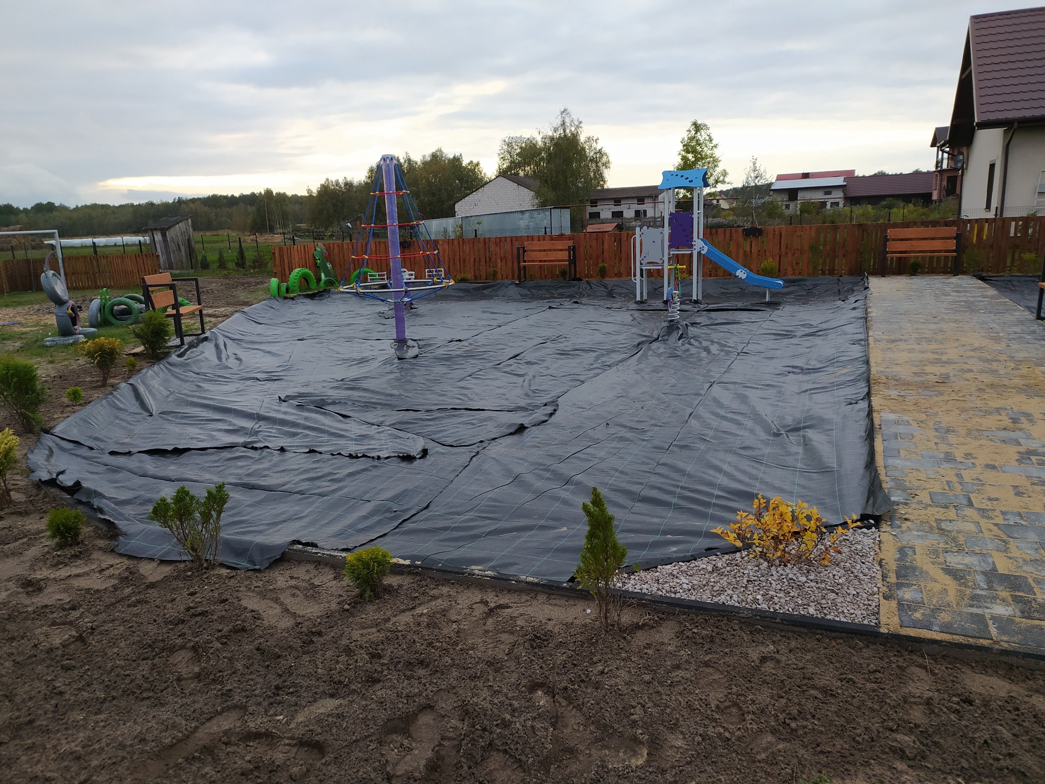 Przygotowanie terenu pod plac zabaw: rozłożona czarna agrowłóknina, posadzone małe tuje i krzewy, w tle drewniany płot i elementy placu zabaw.
