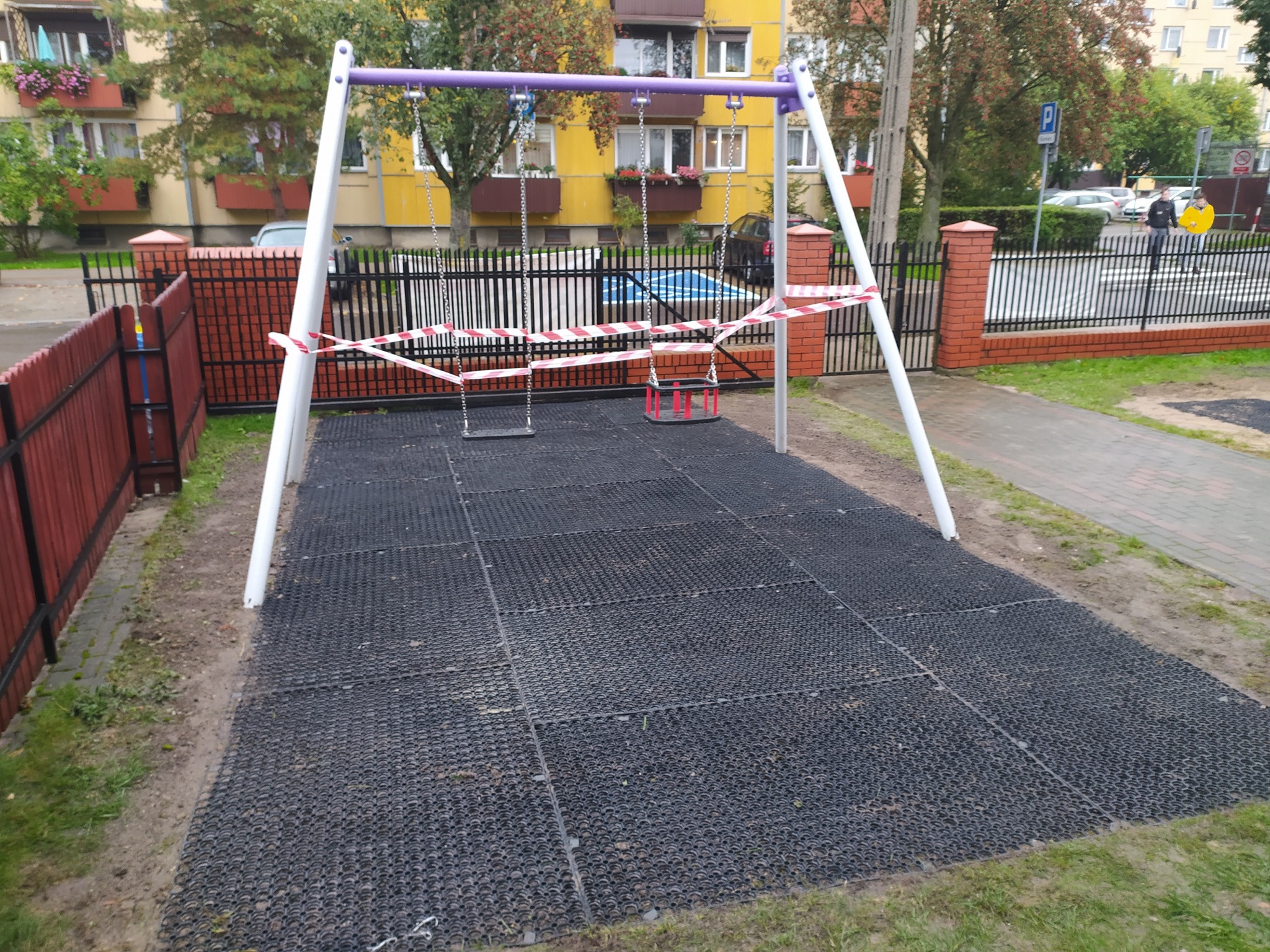Plac zabaw z huśtawkami ogrodzony taśmą ostrzegawczą, na podłożu z gumowych płyt, w tle budynek mieszkalny.