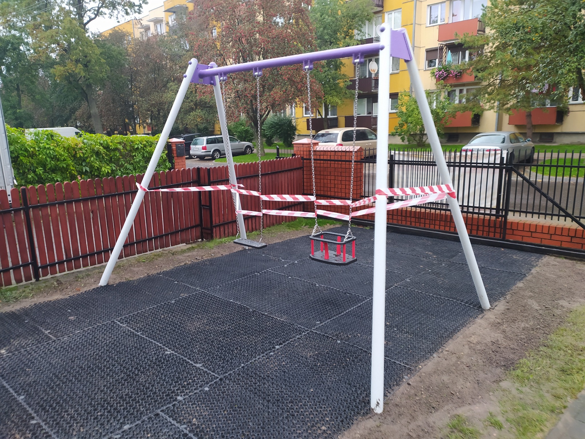 Nowo zainstalowany, fioletowo-biały plac zabaw z huśtawkami, otoczony taśmą ostrzegawczą, na czarnej, gumowej nawierzchni, z widocznym ogrodzeniem i budynkiem mieszkalnym w tle.