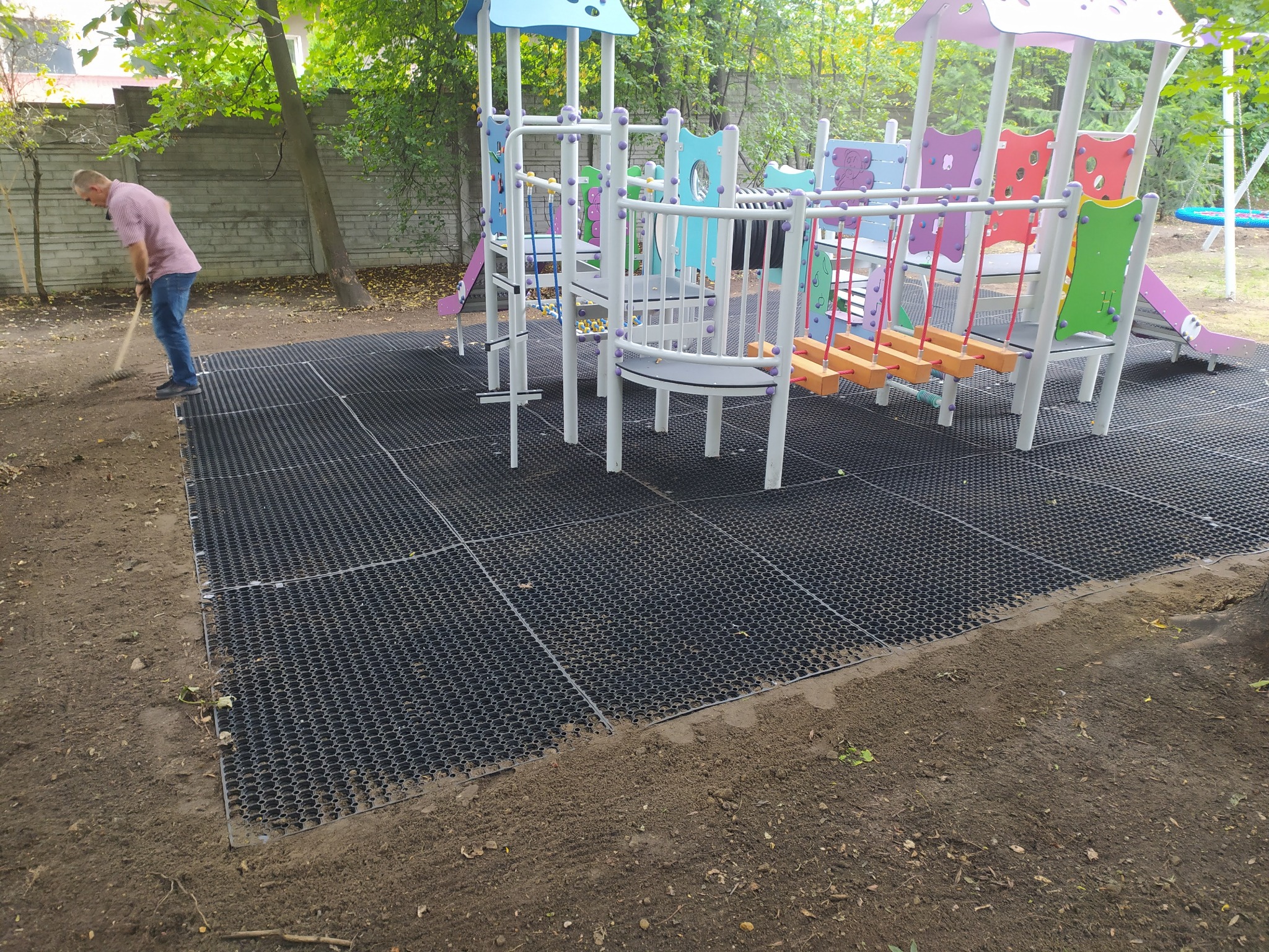 Instalacja czarnej, ażurowej nawierzchni pod plac zabaw z kolorową konstrukcją zabawową w tle, wyrównywanie terenu grabiami.