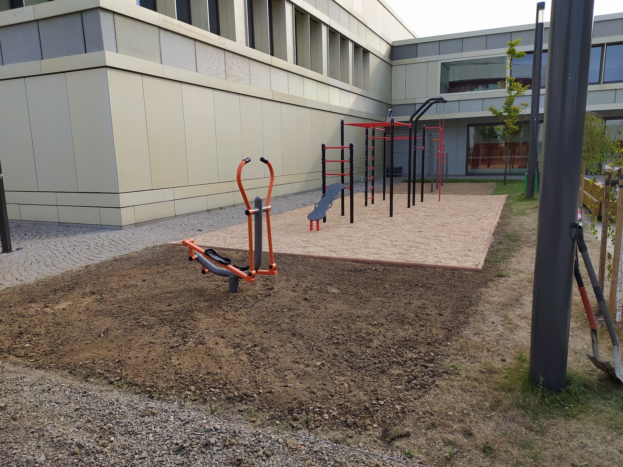 Teren przygotowany pod montaż urządzeń na placu zabaw, widoczne wykopane podłoże oraz urządzenia do ćwiczeń na piasku, w tle budynek.