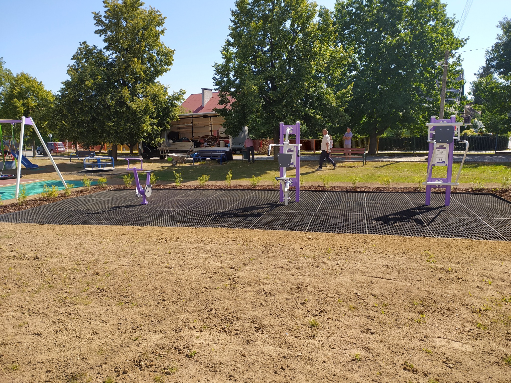 Nowo zainstalowane, fioletowe urządzenia do ćwiczeń na świeżym powietrzu na placu zabaw, z widocznym przygotowanym podłożem i elementami zieleni w tle.
