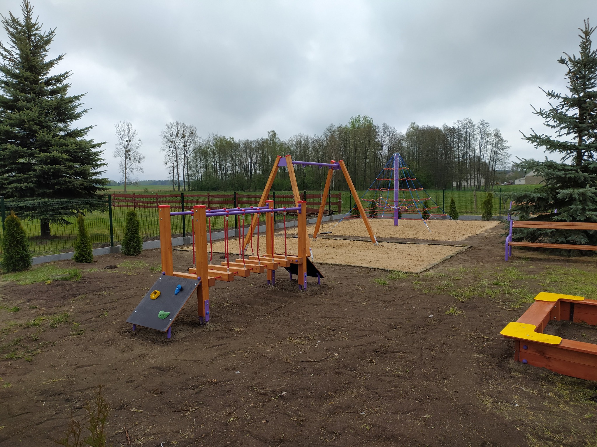 Nowo wybudowany plac zabaw z drewnianymi konstrukcjami, piaskownicą i drabinkami linowymi, otoczony zielenią i ogrodzeniem.