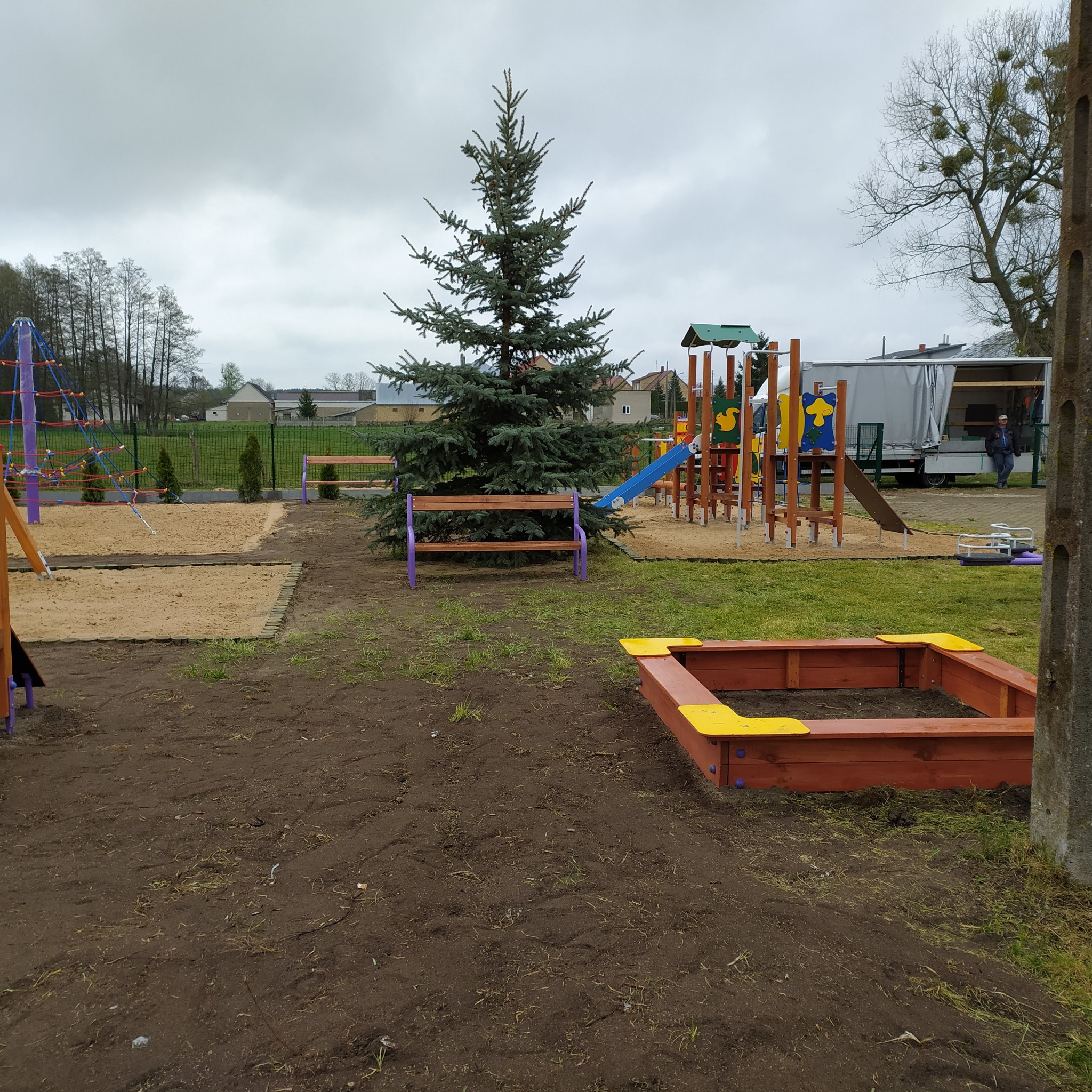 Nowo wybudowany plac zabaw z piaskownicami, drabinkami i zjeżdżalniami, otoczony świeżo przekopaną ziemią i młodymi drzewkami.