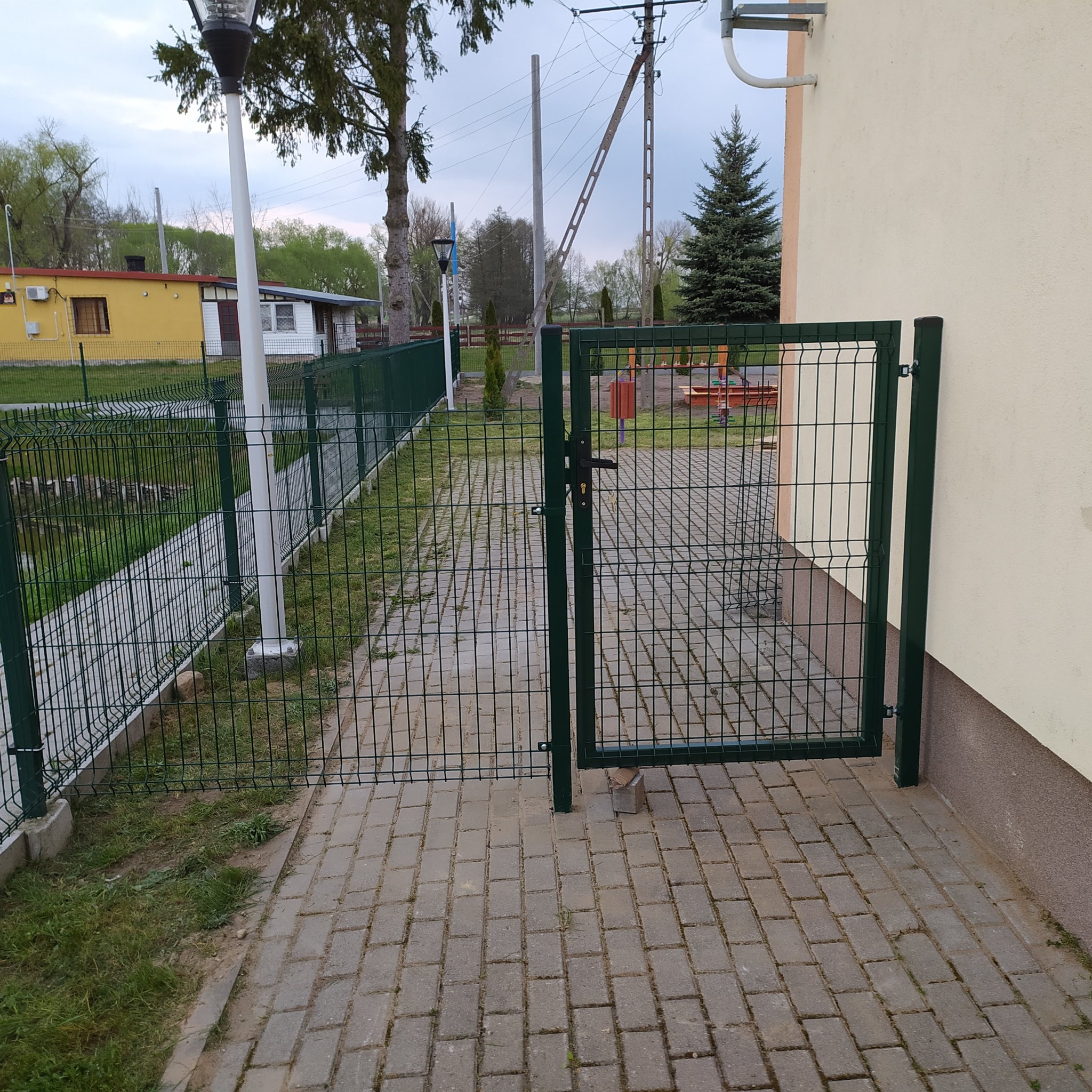 Otwarta, zielona, metalowa furtka z zamkiem w ogrodzeniu z siatki, prowadząca na chodnik z kostki brukowej, widok na posesję z budynkami i drzewami.