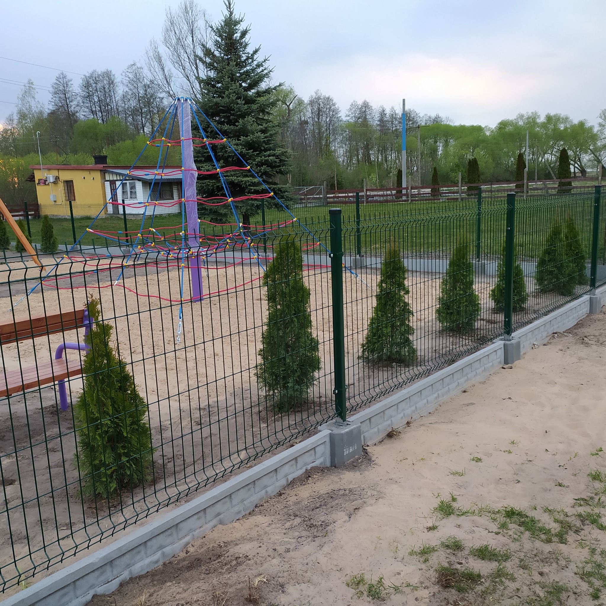 Nowo postawione ogrodzenie panelowe z podmurówką na placu zabaw z piaskiem, z posadzonymi tujami wzdłuż ogrodzenia.