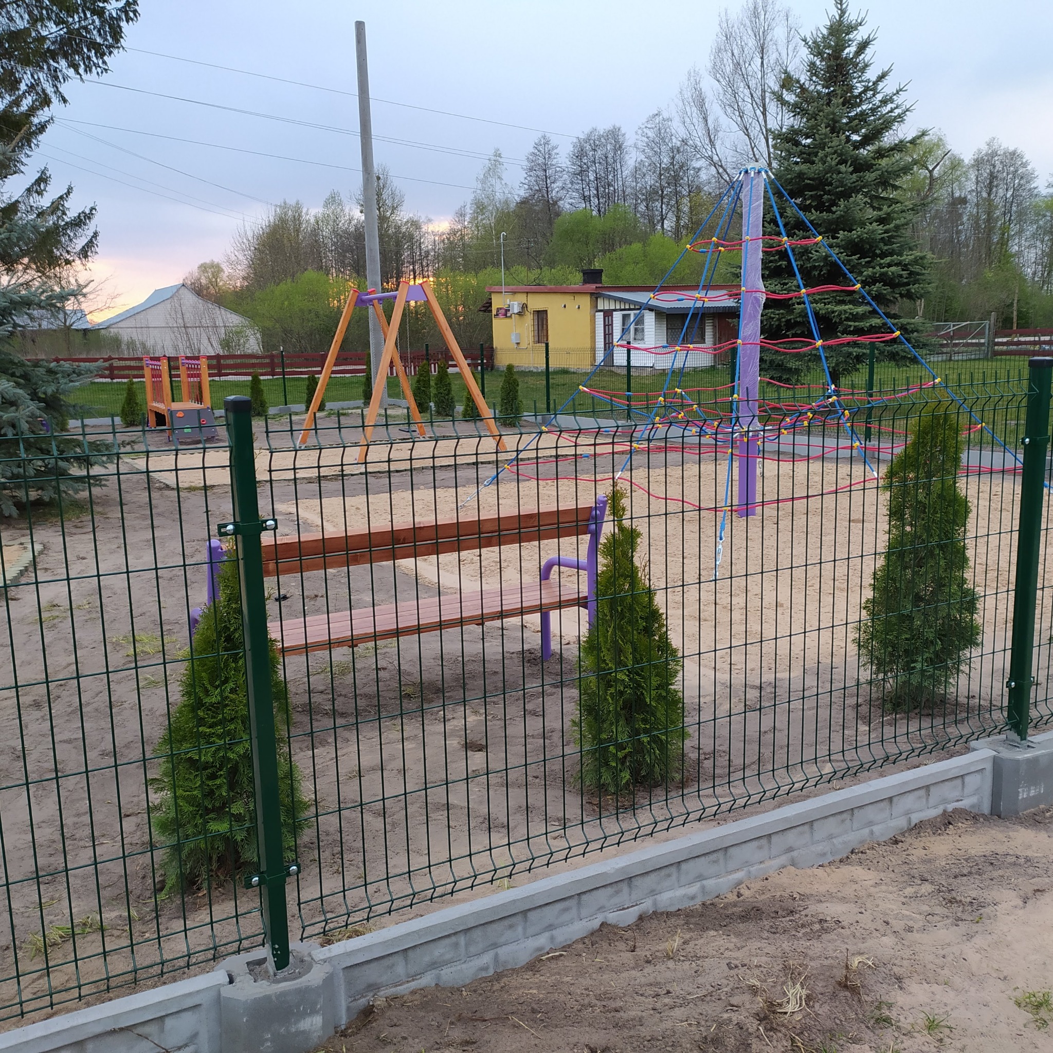 Nowo wybudowany plac zabaw z piaskiem, huśtawkami, piramidą linową i ławką, otoczony zieloną siatką i młodymi tujami, w tle budynki.