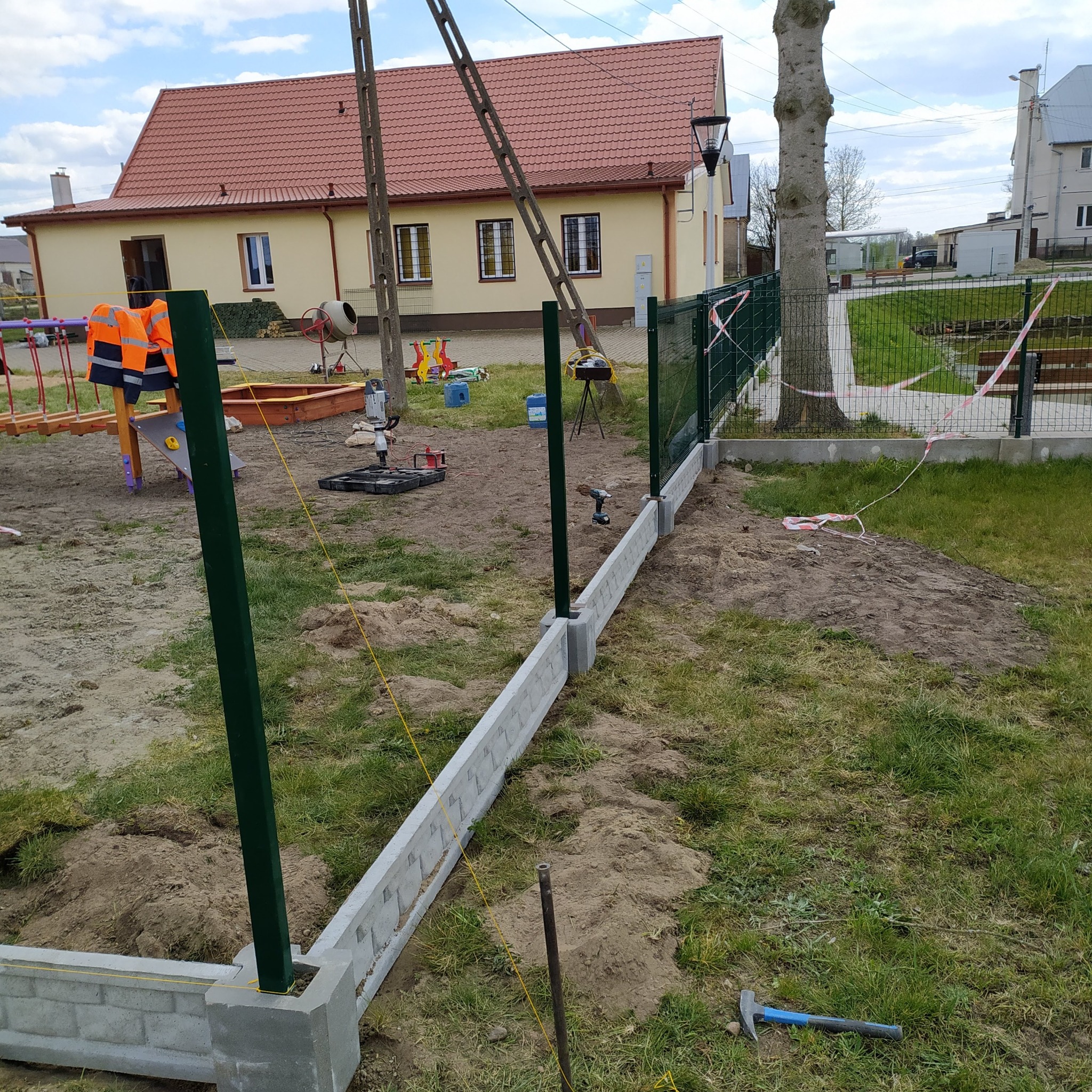 Montaż ogrodzenia panelowego na betonowej podmurówce z użyciem wiertarki akumulatorowej, widoczne słupki w kolorze ciemnej zieleni i narzędzia na trawie.