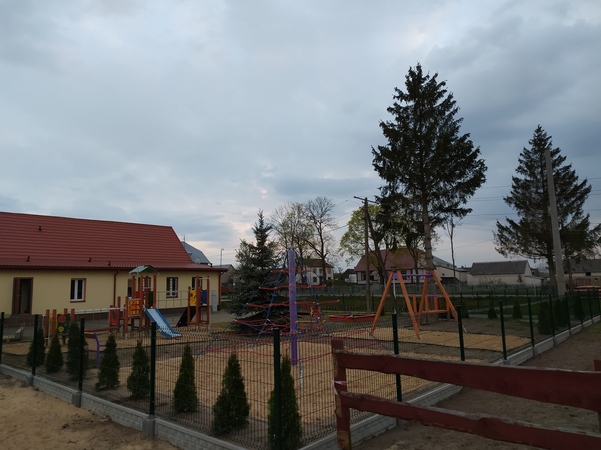 Nowo wybudowany plac zabaw z kolorowymi elementami i piaskiem, otoczony zielonymi choinkami i ogrodzeniem, na tle budynku z czerwoną dachówką i pochmurnego nieba.