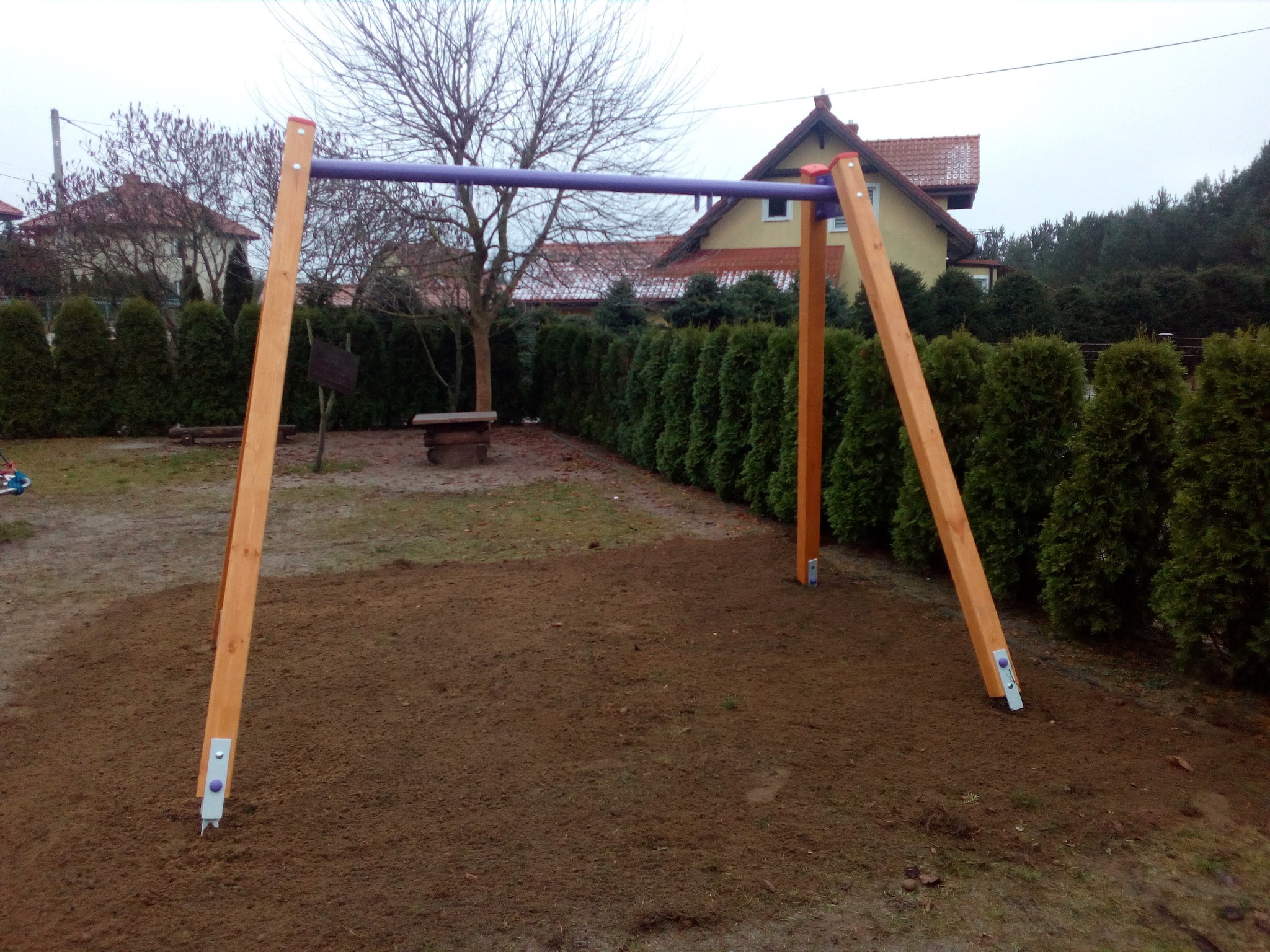 Drewniana konstrukcja huśtawki z fioletowym drążkiem, zamontowana na wyrównanym, brązowym gruncie w ogrodzie z tujami i domem w tle.