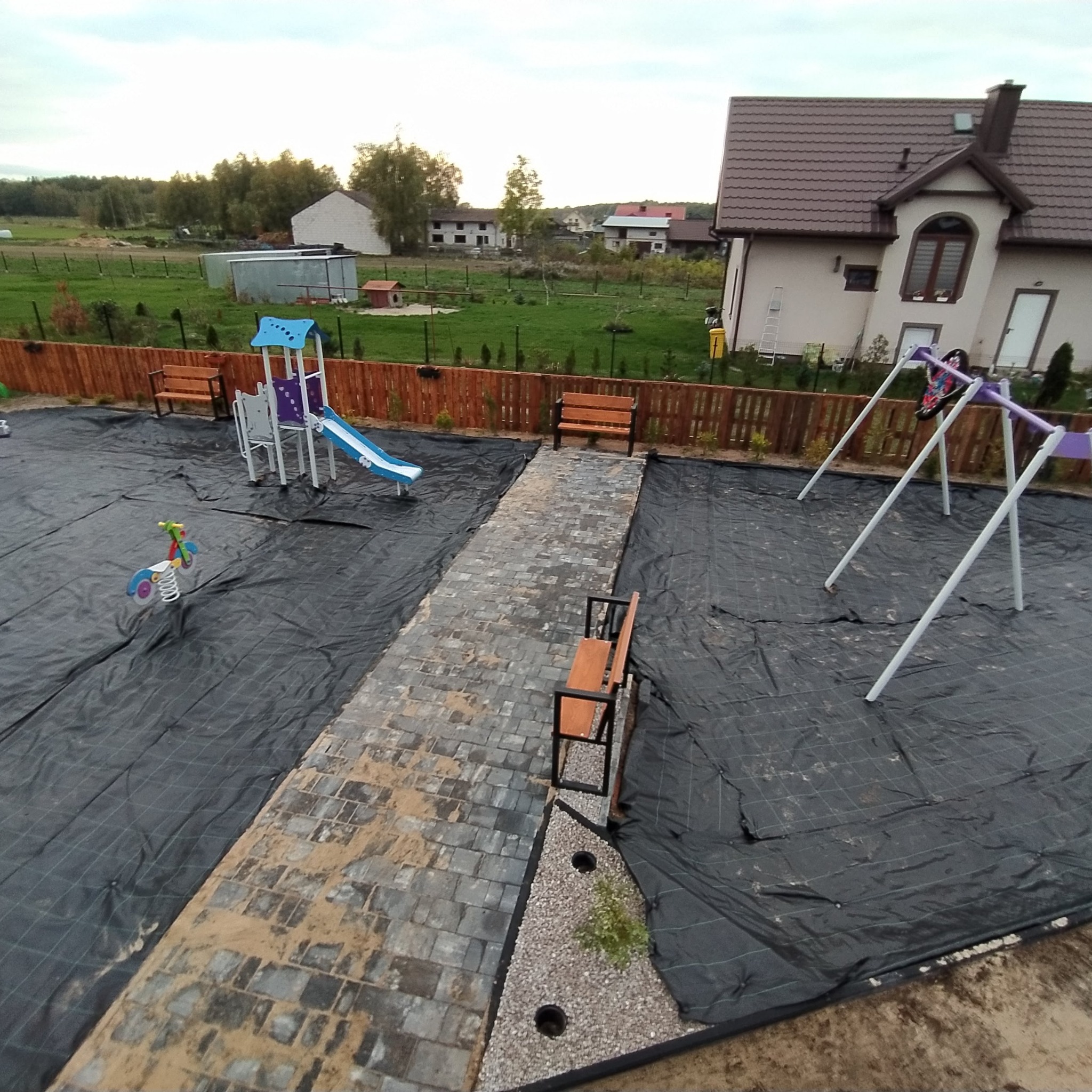Plac zabaw w trakcie budowy: widoczna czarna agrowłóknina, ścieżka z kostki brukowej w trakcie układania, huśtawka, zjeżdżalnia i bujak na sprężynie. W tle dom jednorodzinny i ogrodzenie.