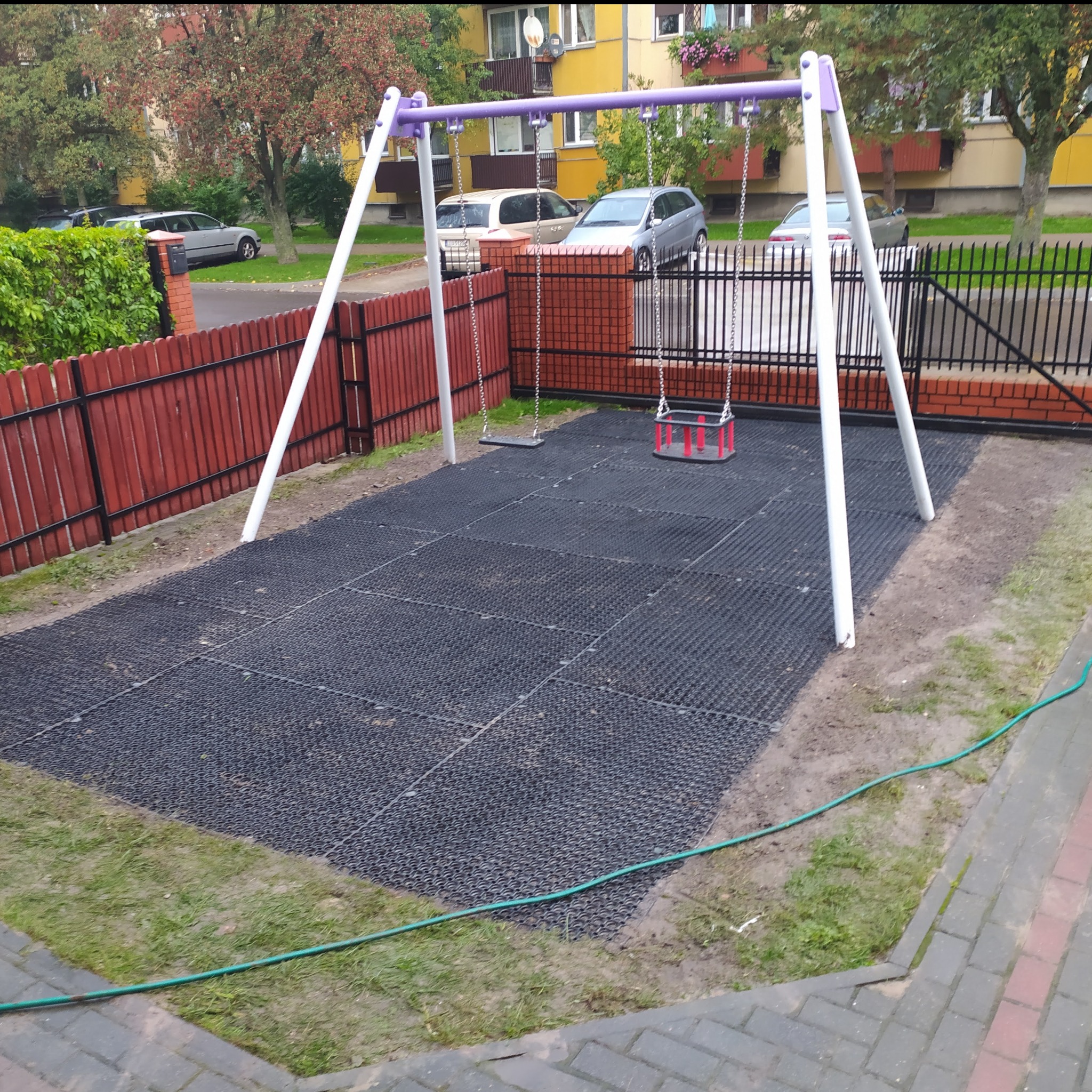 Plac zabaw z huśtawkami na gumowej nawierzchni, otoczony płotem i zielenią przy bloku mieszkalnym.