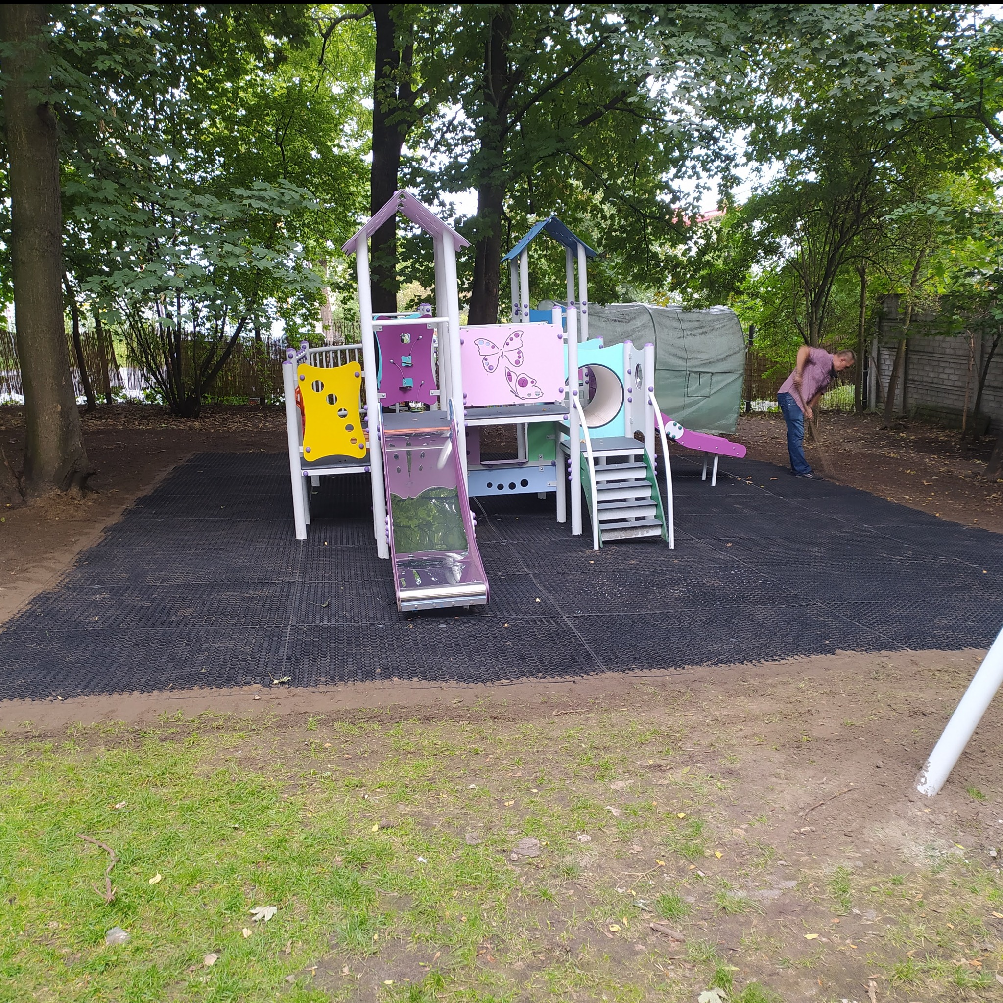 Plac zabaw z kolorową konstrukcją dla dzieci, wyłożony czarną nawierzchnią, mężczyzna zamiatający liście w tle.
