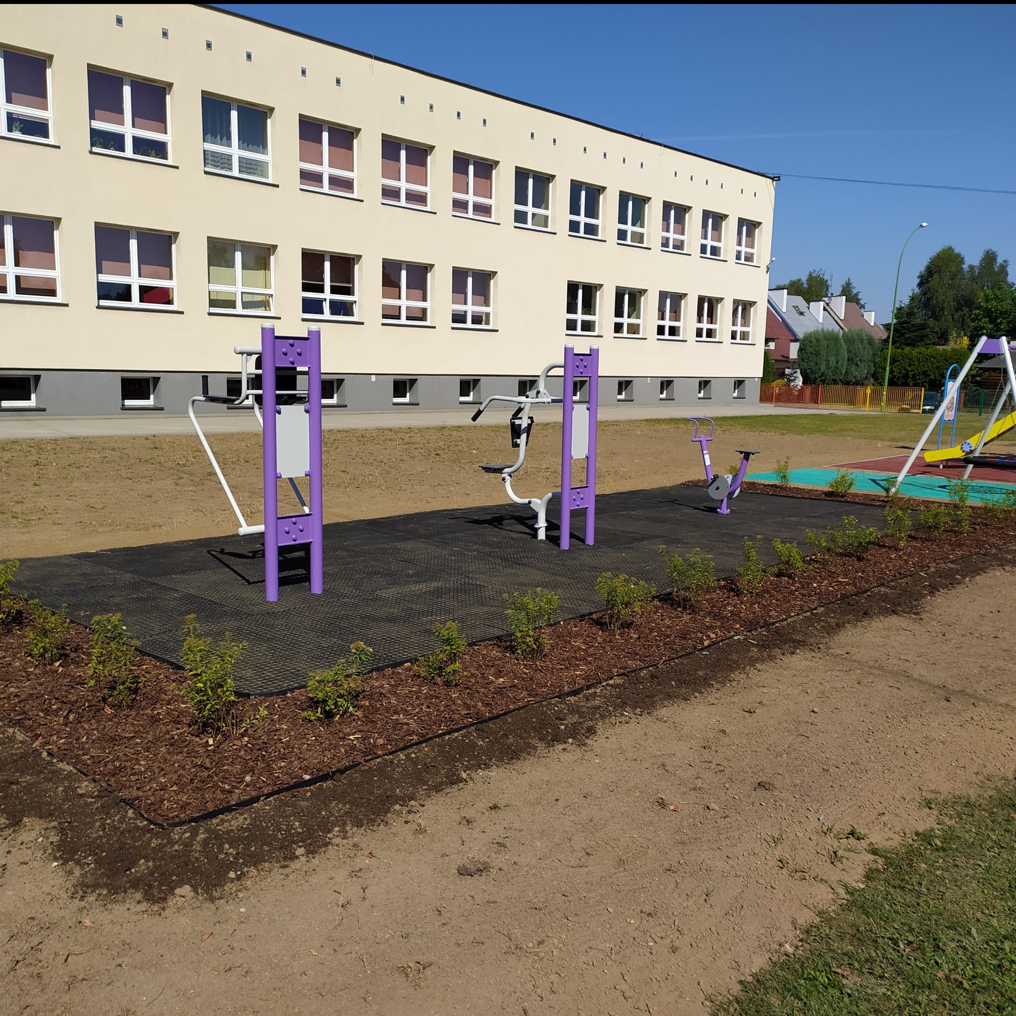 Zewnętrzna siłownia z fioletowymi urządzeniami do ćwiczeń na czarnej, gumowej nawierzchni, obok budynku szkolnego z licznymi oknami.