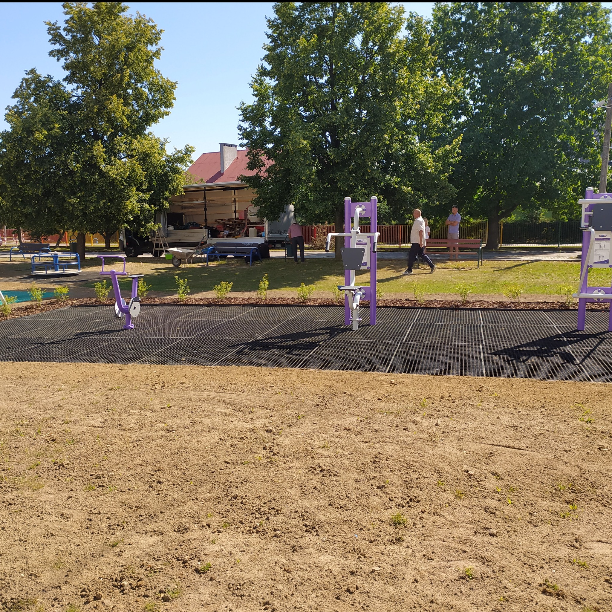 Nowo zainstalowane, fioletowe urządzenia do ćwiczeń na świeżym powietrzu na placu zabaw z czarną, gumową nawierzchnią, otoczone świeżo posadzoną trawą i krzewami, z widocznym w tle samochodem...